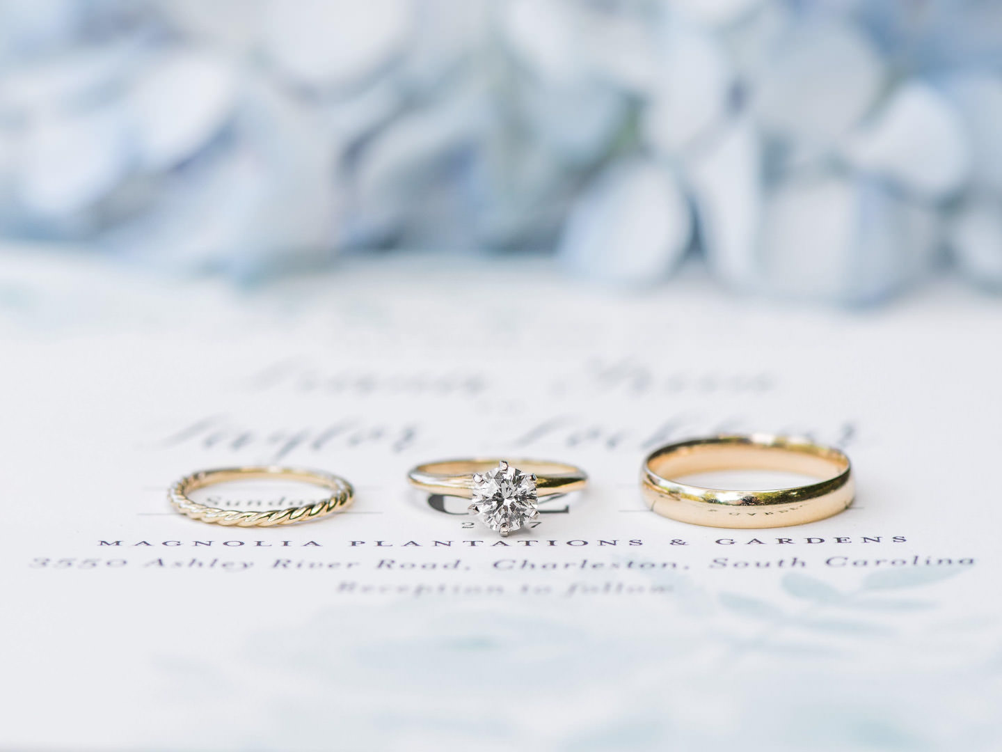 Gold wedding band, gold engagement ring with solitaire, and gold grooms band on invitation suite and hydrangeas